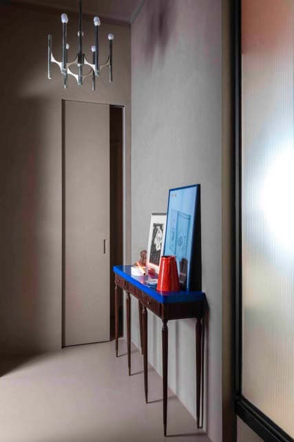 Modern hallway with a blue-topped console table, red vase, and a silver chandelier. Frosted glass door to the right.