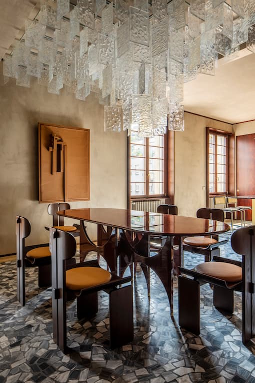 Modern dining room with a glass table, unique wooden chairs, geometric patterned floor, and a striking glass chandelier. Large windows in the background.