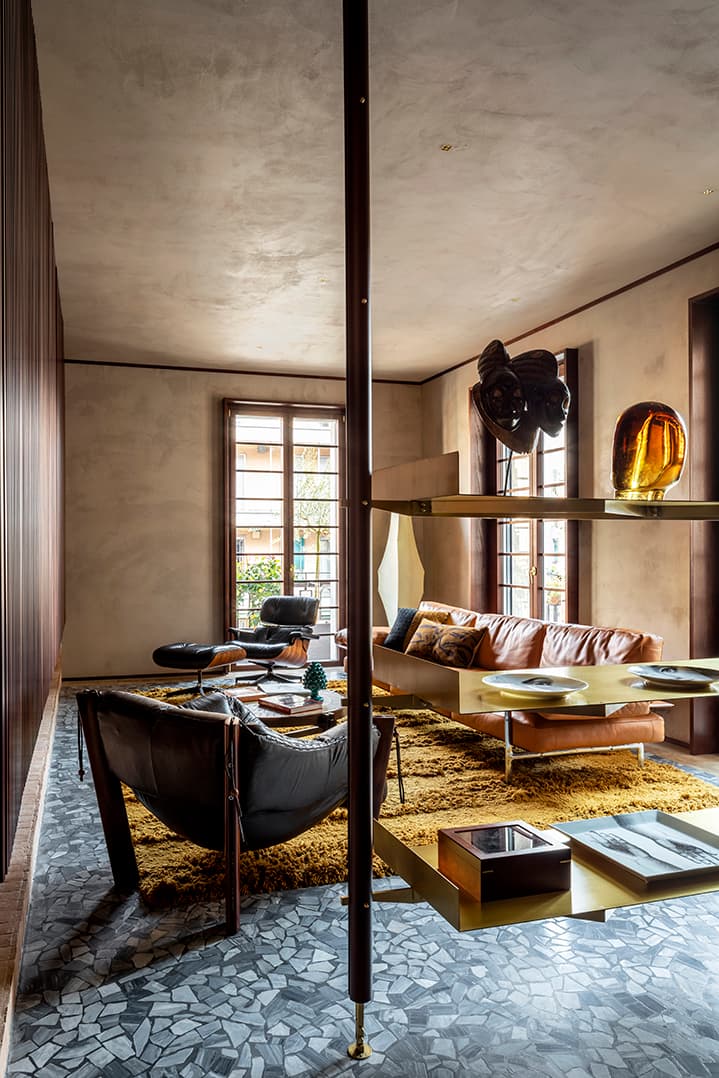 Modern living room with leather sofa, armchairs, and a glass shelving unit. Large windows, textured rug, and terrazzo floor enhance the space.