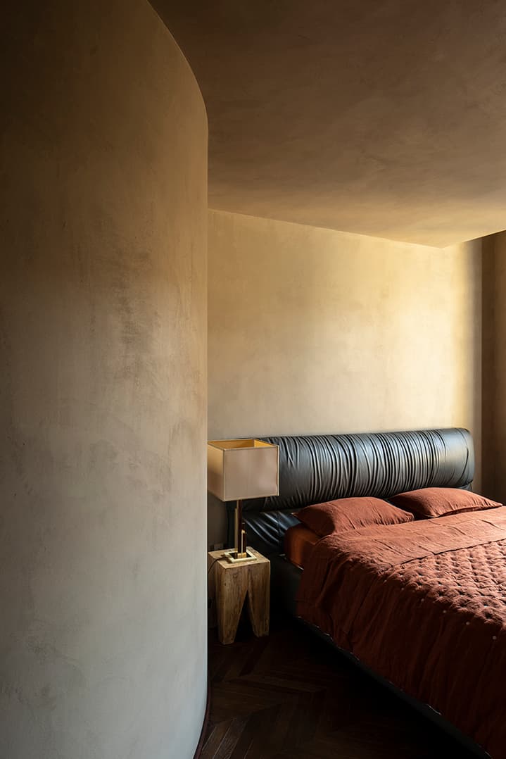 Minimalist bedroom with a curved wall, leather headboard, rust-colored bedding, wooden side table, and a modern lamp.