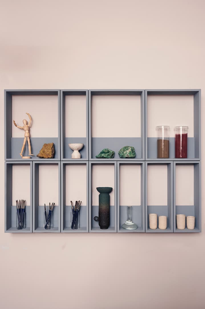 A wall-mounted gray shelf with various items, including a wooden figure, rocks, jars, and small vases, arranged in different compartments.