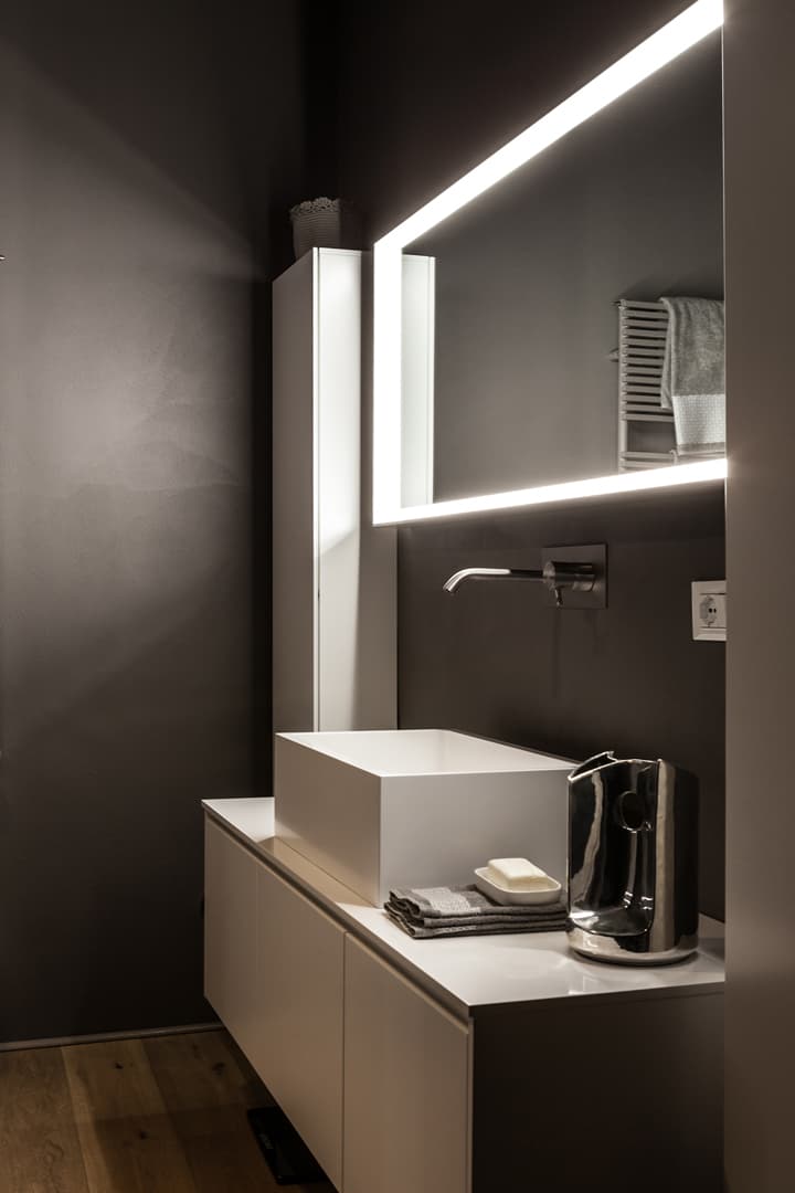 Modern bathroom with a sleek white vanity, square sink, illuminated mirror, and dark walls. A towel and soap are neatly placed on the counter.