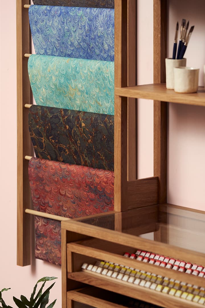 A wooden rack displaying colorful marbled paper sheets, with art supplies like brushes and paints on nearby shelves.
