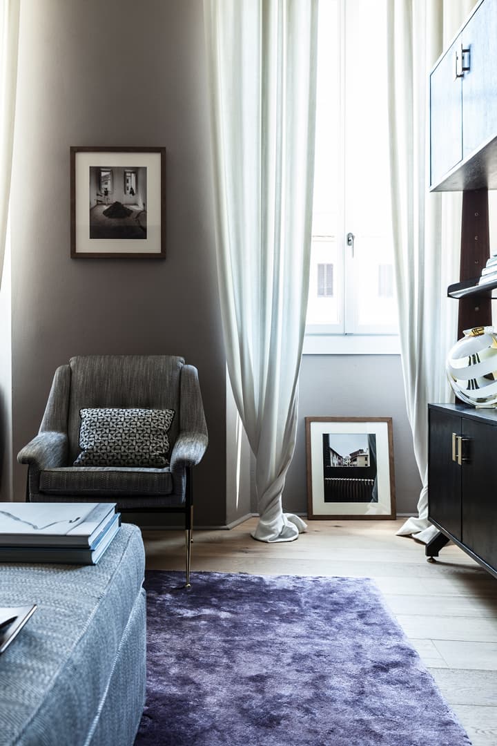 Cozy room with a gray armchair, patterned pillow, purple rug, framed photos, and sheer curtains by a window.