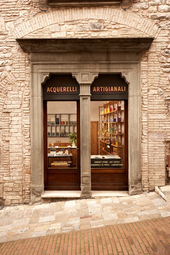 Exterior of a rustic art supply shop with a stone facade, displaying colorful art materials through large windows.