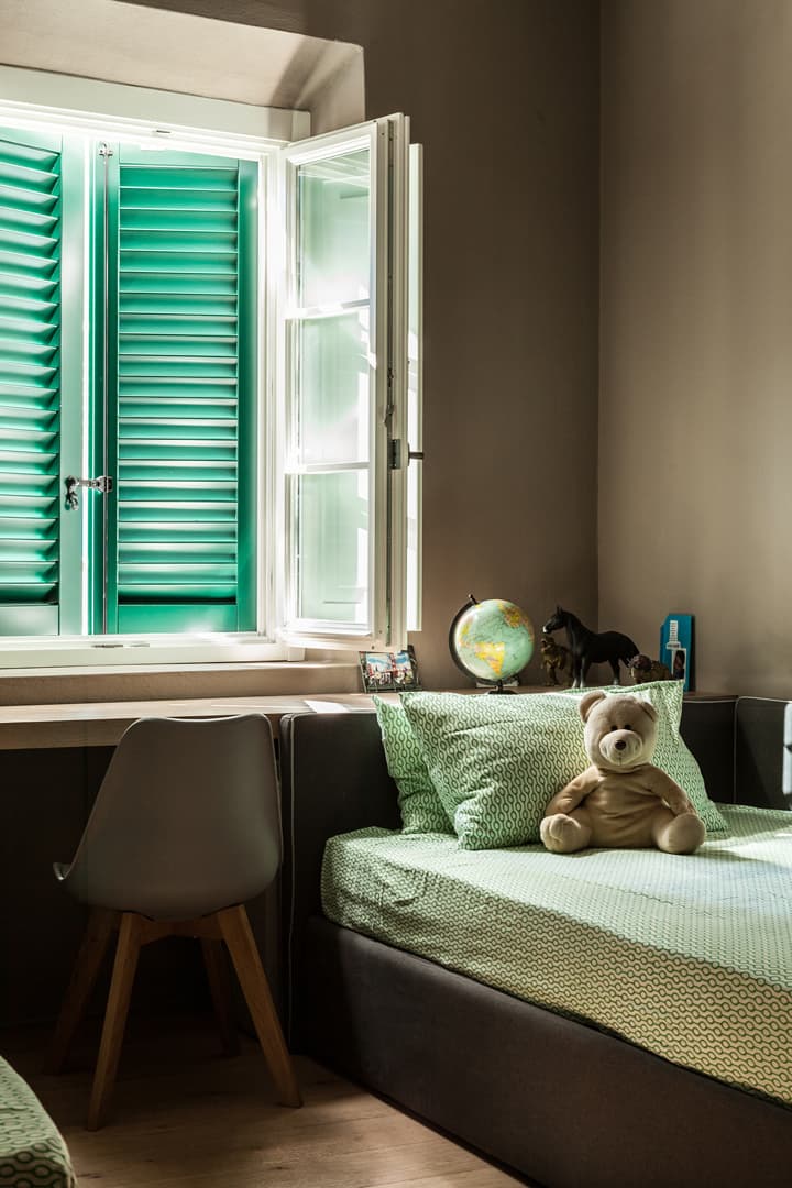 Cozy bedroom with a bed and a globe on a desk by an open window with green shutters, allowing sunlight to stream in.
