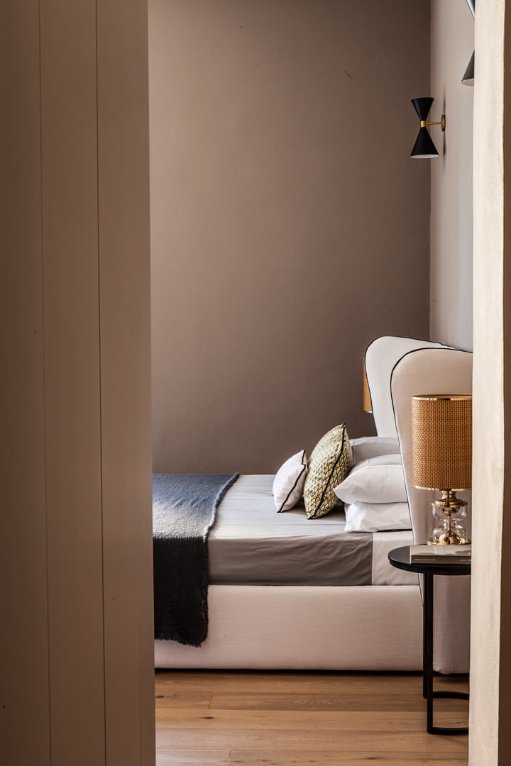 A cozy bedroom with a white bed, gray blanket, patterned pillows, and a side table with a lamp. The walls are painted in a warm, neutral tone.