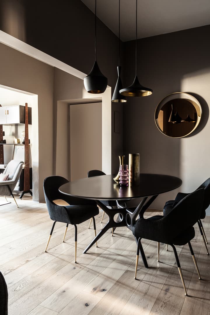 Modern dining room with a round black table, six dark chairs, pendant lights, and a circular mirror on a taupe wall. Light wood flooring.